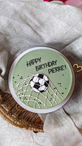 Round green card with soccer ball and net design, featuring 'Happy Birthday Pierre!' text, on a textured fabric background.